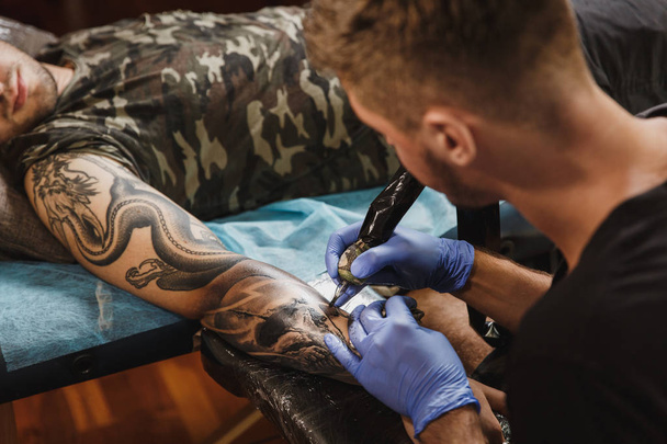 Professional tattooer artist doing picture on hand of man by machine black ink from a jar. Tattoo art on body. Equipment for making tattoo art. Master makes tattooed in light studio. - Photo, Image