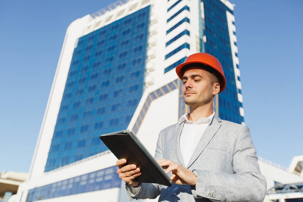 Succesvolle zakenman in grijs pak en beschermende constructie oranje helm met tablet op de achtergrond van het gebouw in blauw-witte kleur. Ingenieur in de helm voor de veiligheid van de werknemers op kantoorgebouwen. - Foto, afbeelding