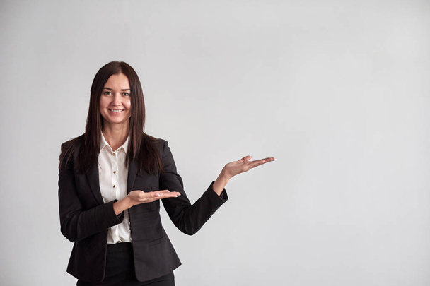 Smiling young businesswoman, showing something, some product or blank copyspace area for advertise slogan or text message, on a gray wall in the office - Photo, image