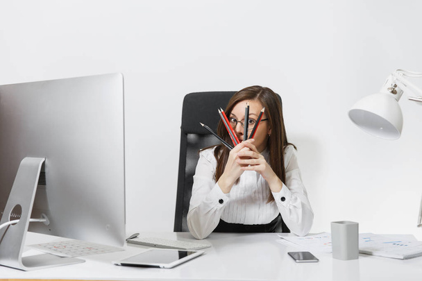 Mooie lachende brown-haar zakenvrouw in pak en bril zitten aan de balie, werken op computer met moderne monitor met documenten in lichte office, verstopt achter potloden op witte achtergrond - Foto, afbeelding