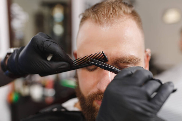Male professional hairdresser serving client, shearing eyebrows with scissors. Ginger stylish young man with thick big beard and short hair getting trendy haircut in black cape. Light barber shop room - Foto, Imagem