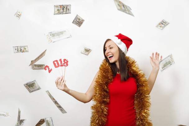 Docela kavkazské šťastná dívka v červených šatech, pozlátko a vánoční čepice drží číslo karty 2018 a peníze bankovky na bílém pozadí. Santa dívka s hotovostí, samostatný. Nový rok dovolená 2018 concept - Fotografie, Obrázek