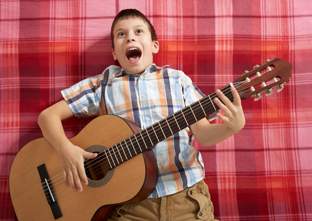 niño tocando música en la guitarra, se encuentra en una manta a cuadros rojo, vista superior
 - Foto, imagen