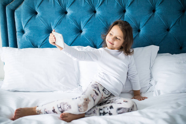 Piękna, uśmiechnięta dziewczynka biorąc selfie na smartfonie leżąc w łóżku miękkie - Zdjęcie, obraz