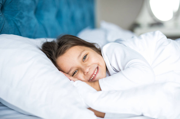 Close-up portrait of laughing little girl lying on bed with hand under pillow, looking at camera and smile. Good morning. - Foto, afbeelding