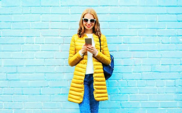 Fashion smiling woman is using smartphone on a blue brick backgr - Zdjęcie, obraz