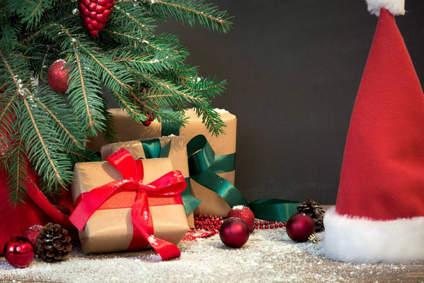 Christmas holiday background. Gifts with a red ribbon, Santa's hat and decor under a Christmas tree on a wooden board. Copy space on chalkboard. - Photo, image