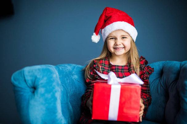 Piękno piękna dziewczyna ubrana w sukienkę i santa hat z Boże Narodzenie prezent siedzący uśmiechający się w fotelu w domu - Zdjęcie, obraz