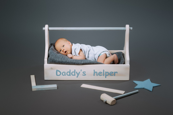 little baby in bodysuit lying in wooden toolkit with daddys helper lettering and hammer near by - Foto, afbeelding