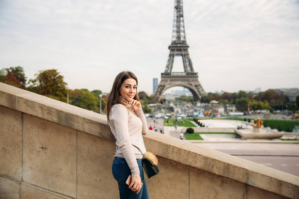 Beautiful girl posing to the photographer against the background of the Eiffel Tower. Autumn photosession. Sunny weather. Beautiful smile and makeup - Photo, image