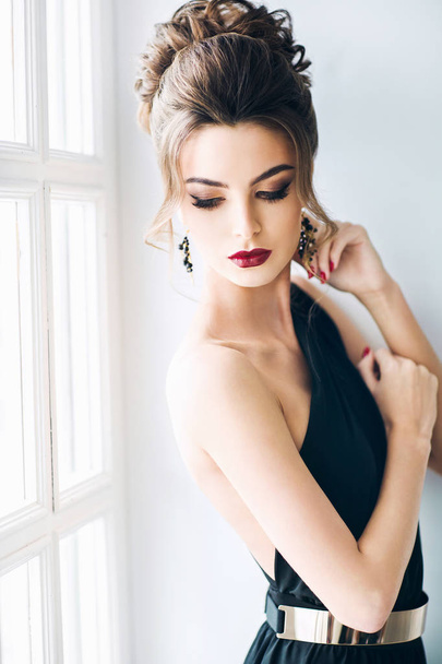 beautiful woman with hairstyle in elegant black dress near window - Zdjęcie, obraz