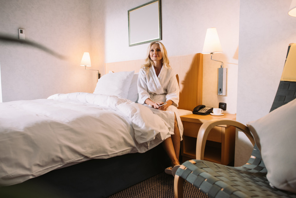 érett nő kezében ágyon ül, és keres el szállodai szobában smartphone - Fotó, kép