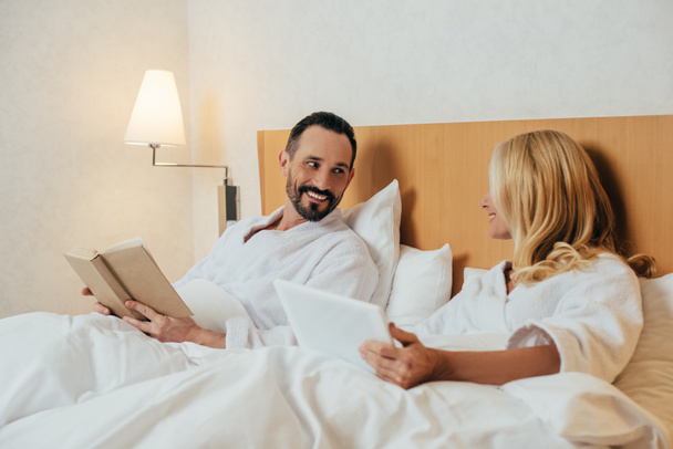 středního věku pár s digitálním tabletu a knihy se navzájem při ležení v posteli v hotelovém pokoji - Fotografie, Obrázek