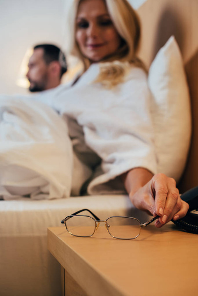 close-up view of woman putting eyeglasses on table while lying with husband in bed at hotel room - Foto, afbeelding