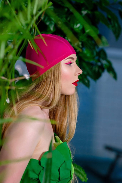 Портрет спортивной красивой блондинки с макияжем в плавательной шапке и костюмом в бассейне с зелеными цветами и растениями
 - Фото, изображение