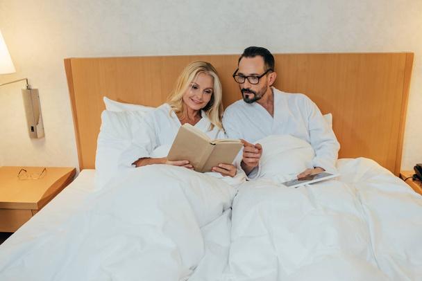 mature couple in bathrobes reading book and using digital tablet in bed in hotel room - Fotografie, Obrázek