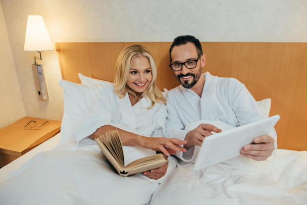 usmíval se dospělý pár čtení knihy a pomocí digitálních tablet zároveň leží vedle sebe v posteli v hotelovém pokoji - Fotografie, Obrázek