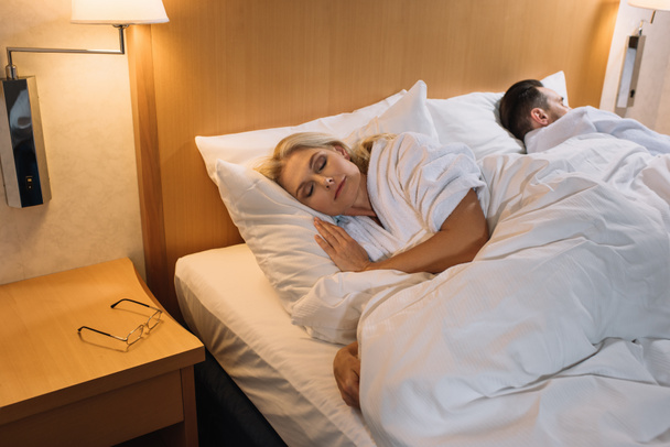 mature couple in bathrobes sleeping in bed and eyeglasses on table in hotel room  - Foto, Imagen