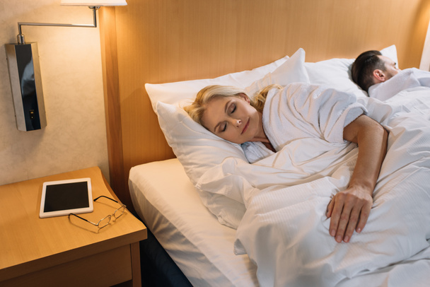 kypsä pari kylpytakit nukkuu sängyssä ja digitaalinen tabletti silmälasit pöydällä hotellihuoneessa
  - Valokuva, kuva