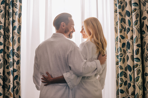 Rückansicht eines verliebten Paares mittleren Alters in Bademänteln und lächelnd im Hotelzimmer  - Foto, Bild