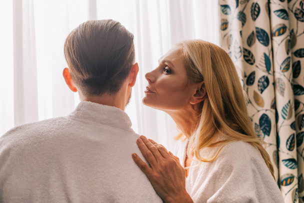 profile view of mature woman able to kiss her husband wearing bathrobe in hotel room - Fotó, kép