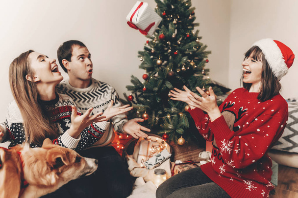 glückliche Freunde in stylischen Pullovern und süßer lustiger Hund, der Geschenke am Weihnachtsbaum mit Lichtern austauscht - Foto, Bild