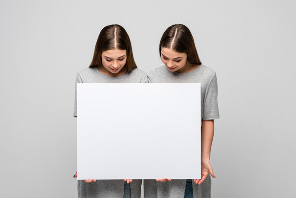 retrato de gemelos jóvenes mirando la pancarta en blanco en las manos aisladas en gris
 - Foto, Imagen