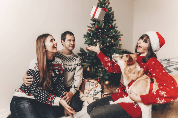 happy friends in stylish sweaters and cute funny dog exchanging gifts sitting at Christmas tree with lights - Photo, Image