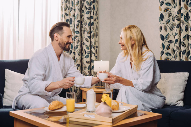 feliz pareja de mediana edad en albornoces beber café durante el desayuno en la habitación del hotel
 - Foto, Imagen