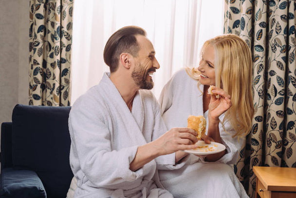 feliz pareja madura en albornoces comer pastelería para el desayuno juntos en la habitación de hotel
  - Foto, Imagen
