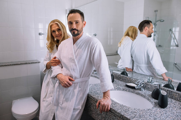 счастливая зрелая пара в халатах улыбается перед камерой в ванной комнате отеля
 - Фото, изображение