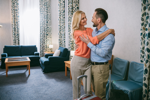 Happy dospělý pár objímání stoje s kufry v hotelovém pokoji - Fotografie, Obrázek