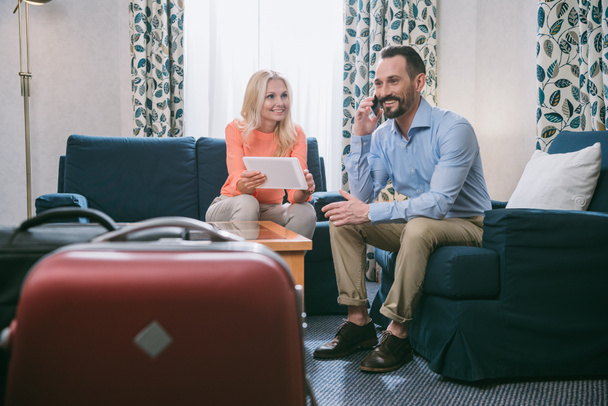 hymyilevä kypsä pari, joka käyttää älypuhelinta ja digitaalista tablettia istuessaan matkalaukut hotellihuoneessa
 - Valokuva, kuva