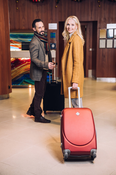 happy mature couple with suitcases standing in hotel hallway - Fotó, kép