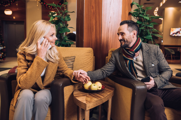 fröhliches reifes Paar, das sich Händchen hält und einander ansieht, während die Frau im Hotel auf dem Smartphone spricht - Foto, Bild