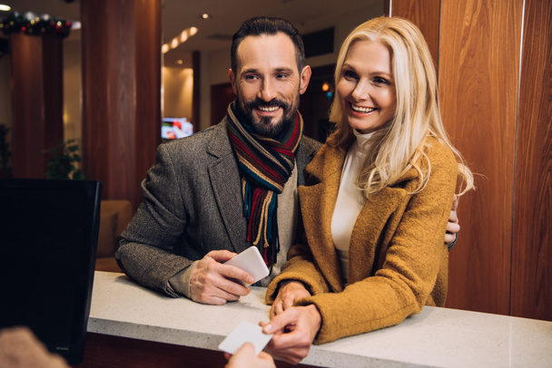 pareja madura sonriente sosteniendo teléfono inteligente y tarjeta en la recepción en el hotel
 - Foto, Imagen
