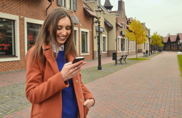 A beautiful girl with brown hair stands on the alley, looks at the phone and smiles. Dressed in a coat, hand in pocket. Around the building and the tree. - Fotoğraf, Görsel