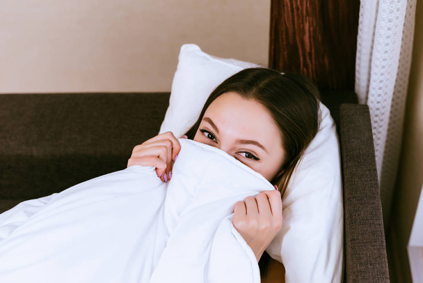 funny young girl lies in bed, covers half of face with white blanket - Foto, immagini
