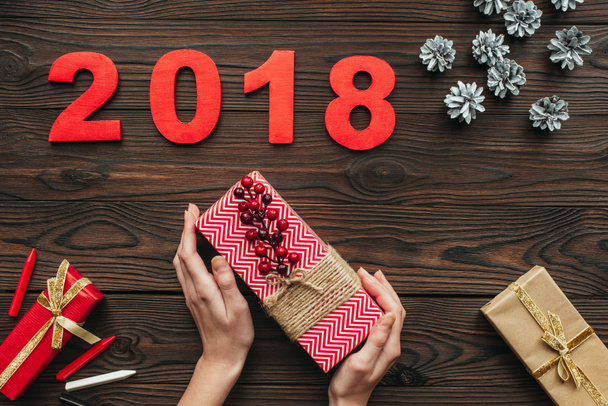cropped shot of woman holding wrapped gift in hands with pine cons and 2018 around on wooden tabletop - Фото, изображение
