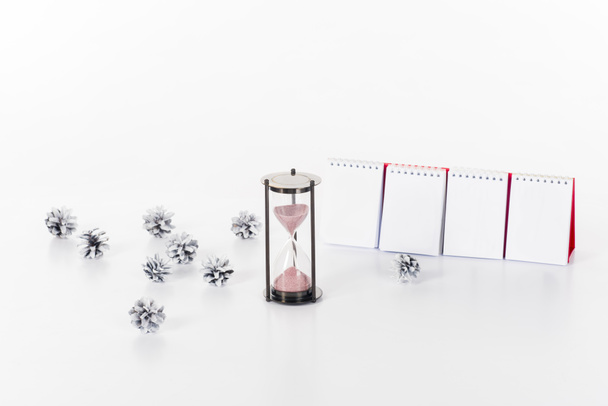 закрывающийся вид чистого календаря, песчаных часов и сосновых шишек, изолированных на белом
 - Фото, изображение