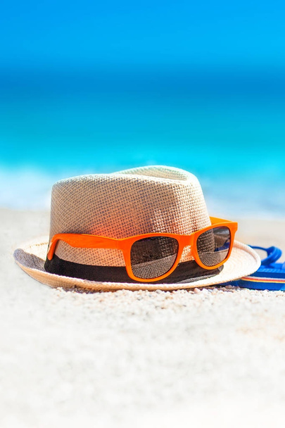 Chanclas, sombrero de playa, gafas de sol en la arena. Concepto vacaciones de verano. Día soleado en la playa tropical con horizonte azul del mar
 - Foto, Imagen