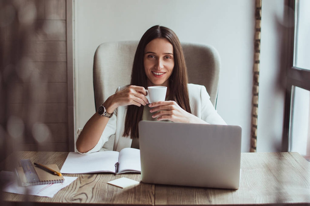 Что может быть лучше свежей чашки кофе? Красивая молодая деловая женщина держит чашку кофе и смотрит в камеру с улыбкой, сидя за рабочим столом
 - Фото, изображение