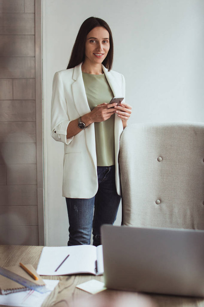 Loving her job. Beautiful young businesswoman using her smart phone and looking at camera with smile while standing at her office desk - Fotoğraf, Görsel