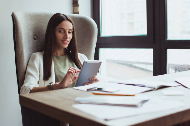 Dia perfeito no cargo. Bela jovem empresária usando seu tablet digital com sorriso enquanto se senta em sua mesa de escritório
 - Foto, Imagem