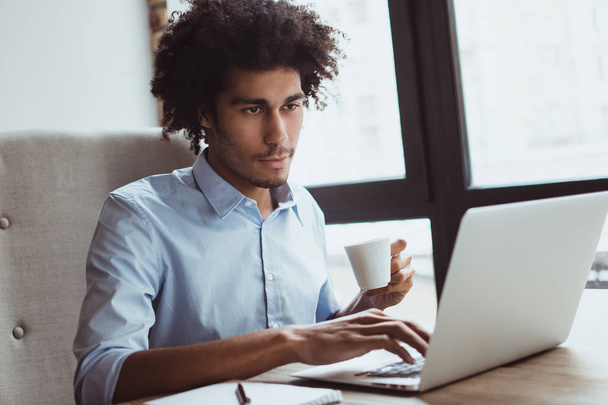Кава і звіт. Красивий молодий бізнесмен використовує свій ноутбук і п'є каву, сидячи за офісним столом
 - Фото, зображення