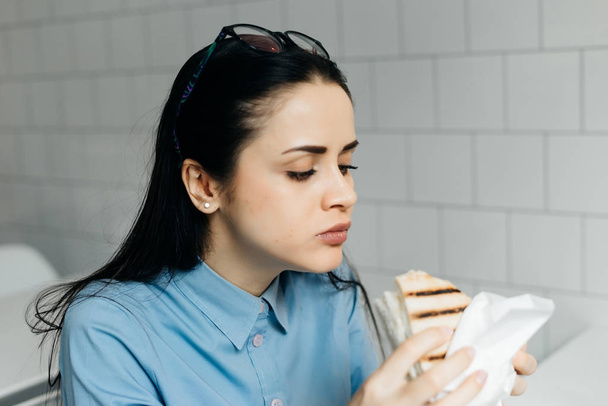 busy young girl student in a blue shirt eating a sandwich in a cafe - Photo, image