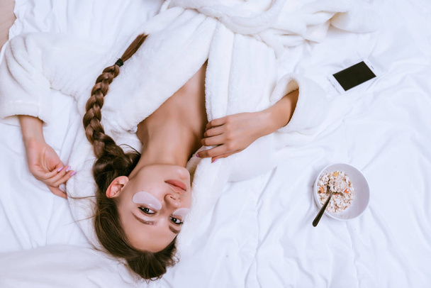 beautiful young girl in a white terry robe lies on the bed, under the eyes of white silicone patches, next to a plate of oatmeal porridge - Fotoğraf, Görsel