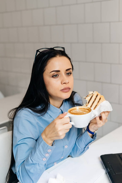 attractive black-haired girl in blue shirt, student, having breakfast in cafe with sandwich and coffee before classes - Photo, Image
