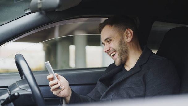 Atractivo hombre de negocios sentado dentro del coche riendo mientras usa el teléfono inteligente después del viaje
 - Foto, Imagen