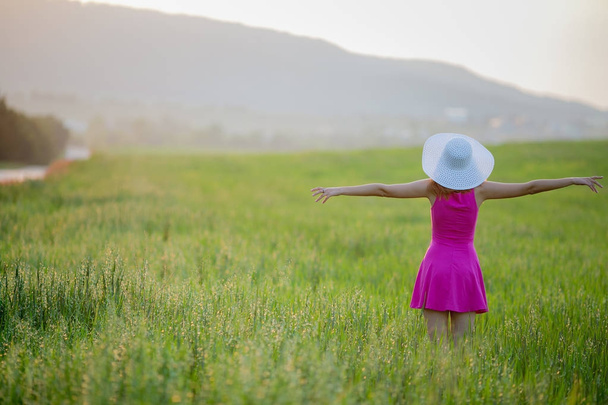 young woman in purple dress and white hat walking on green field with mountains on background, back view - Fotó, kép